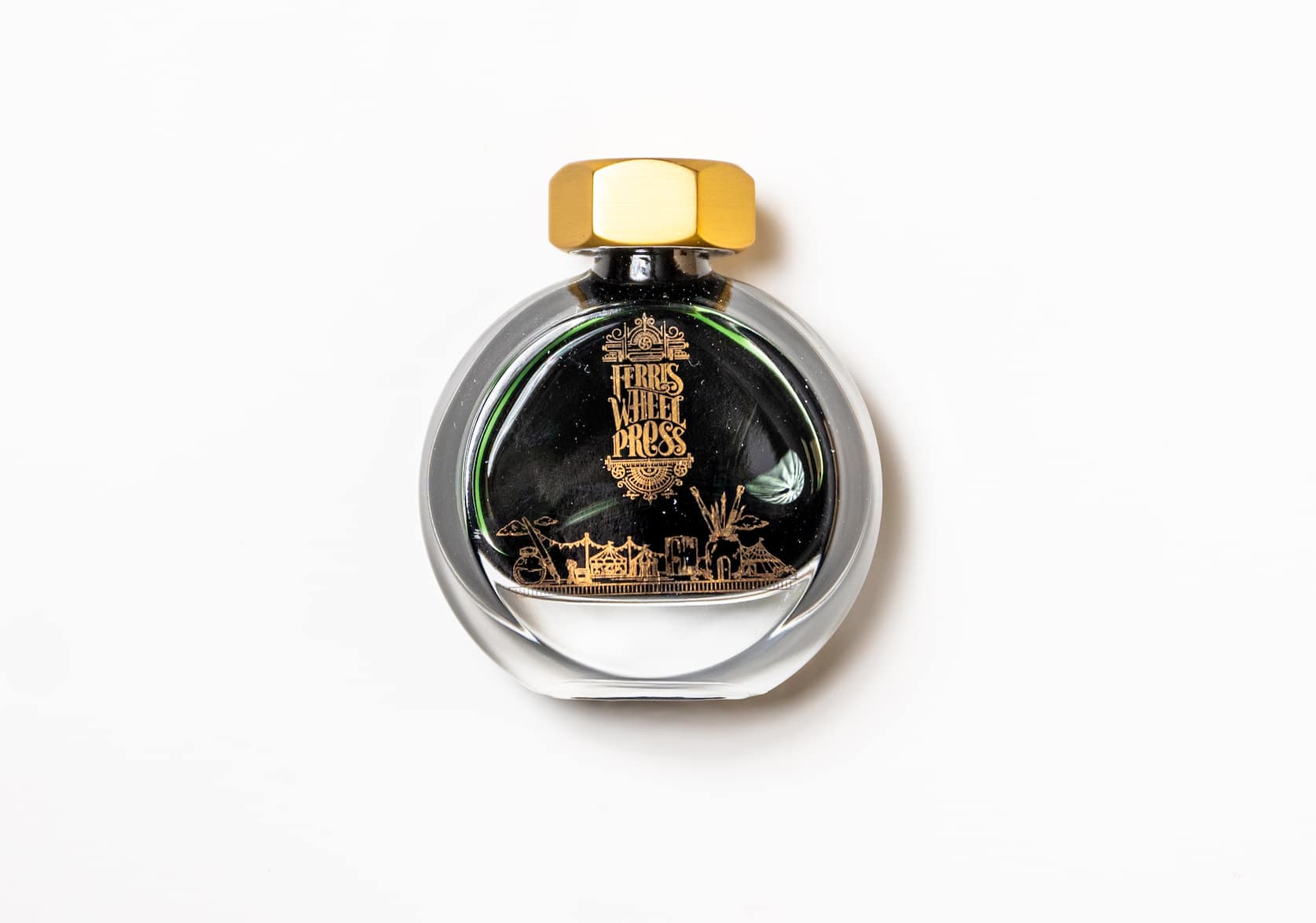 A round glass bottle of ink with a gold lid. On the bottle there is a small gold illustration featuring a merry-go-round and circus tent.