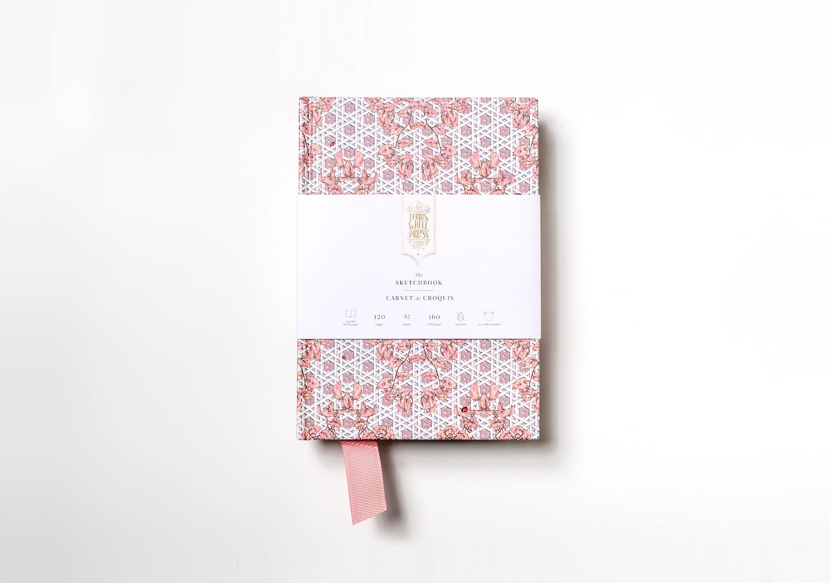 A pink sketchbook covered in paper with a hand-illustrated rattan pattern featuring a pink fabric ribbon sticking out of the bottom. Gold text on the notebook's packaging reads: Ferris Wheel Press logo. The Sketchbook. Carnet de Croquis.