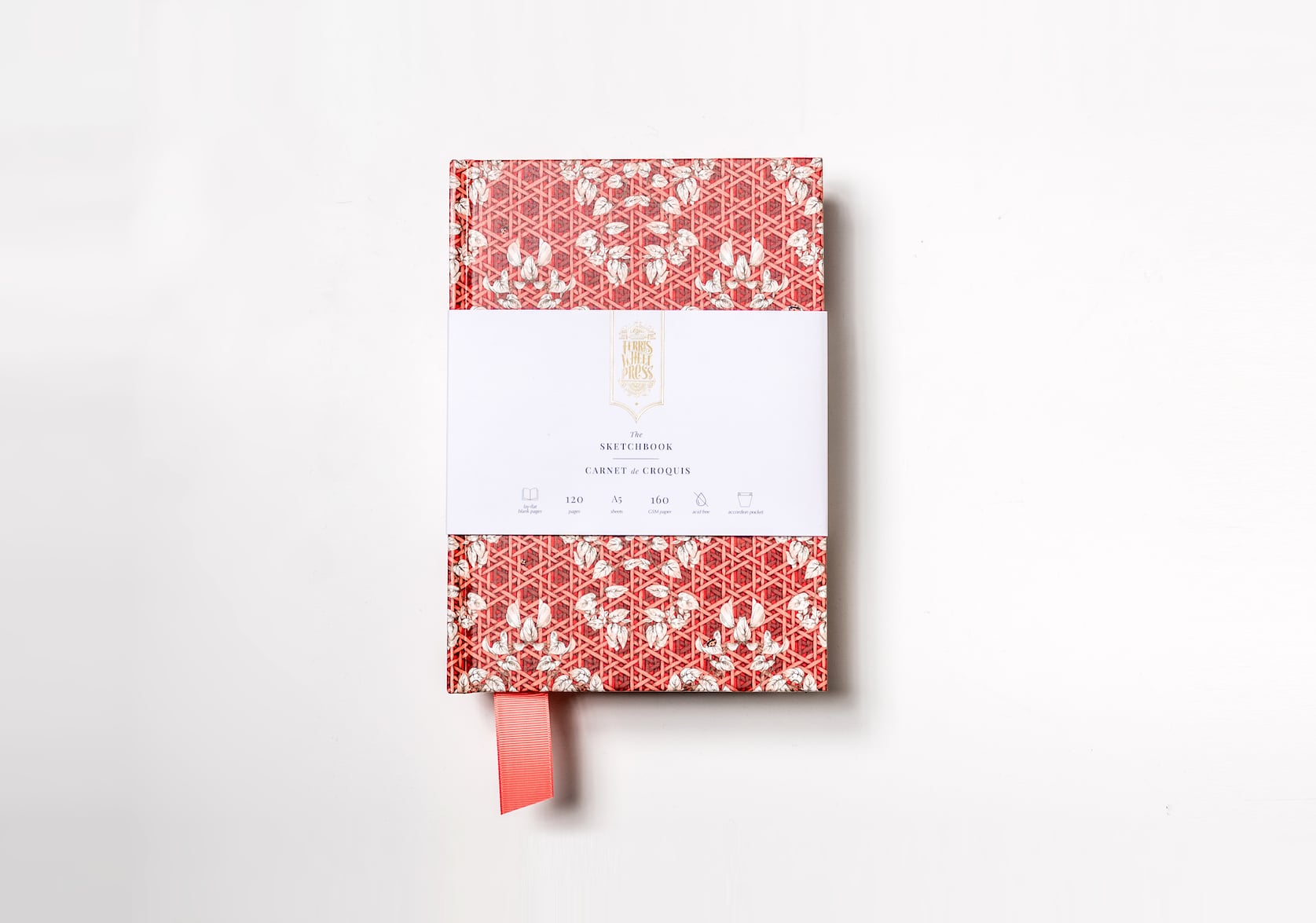 A red sketchbook covered in paper with a hand-illustrated rattan pattern featuring a red fabric ribbon sticking out of the bottom. Gold text on the notebook's packaging reads: Ferris Wheel Press logo. The Sketchbook. Carnet de Croquis.