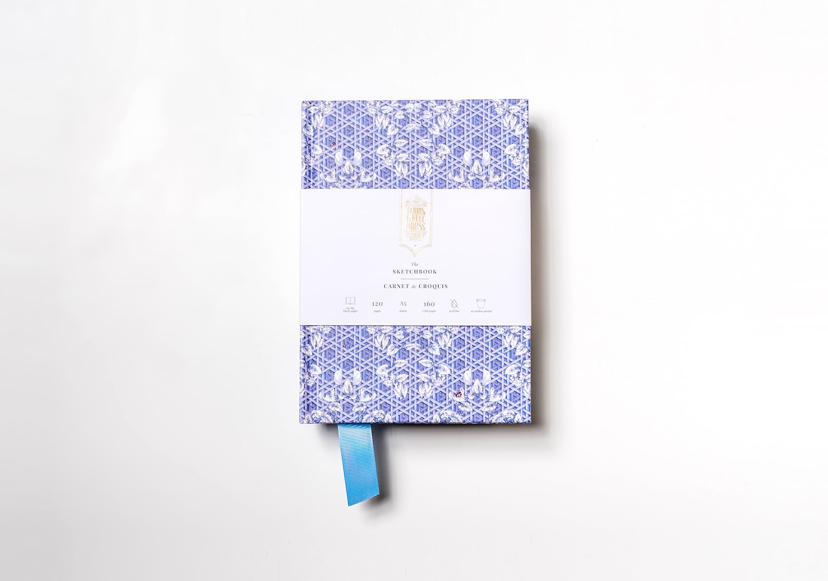 A violet blue sketchbook covered in paper with a hand-illustrated rattan pattern featuring a violet blue fabric ribbon sticking out of the bottom. Gold text on the notebook's packaging reads: Ferris Wheel Press logo. The Sketchbook. Carnet de Croquis.