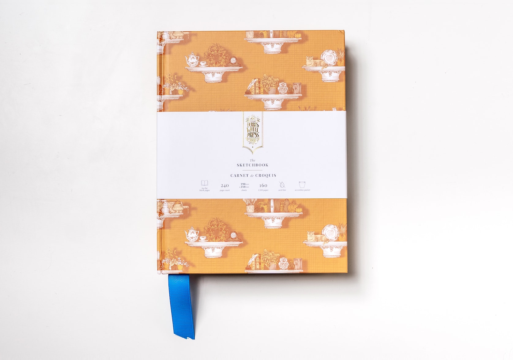 An auburn sketchbook covered in paper with a hand-illustration of white pillar shelving with various objects featuring a teal fabric ribbon sticking out of the bottom. Gold text on the notebook's packaging reads: Ferris Wheel Press logo. The Sketchbook. Carnet de Croquis.