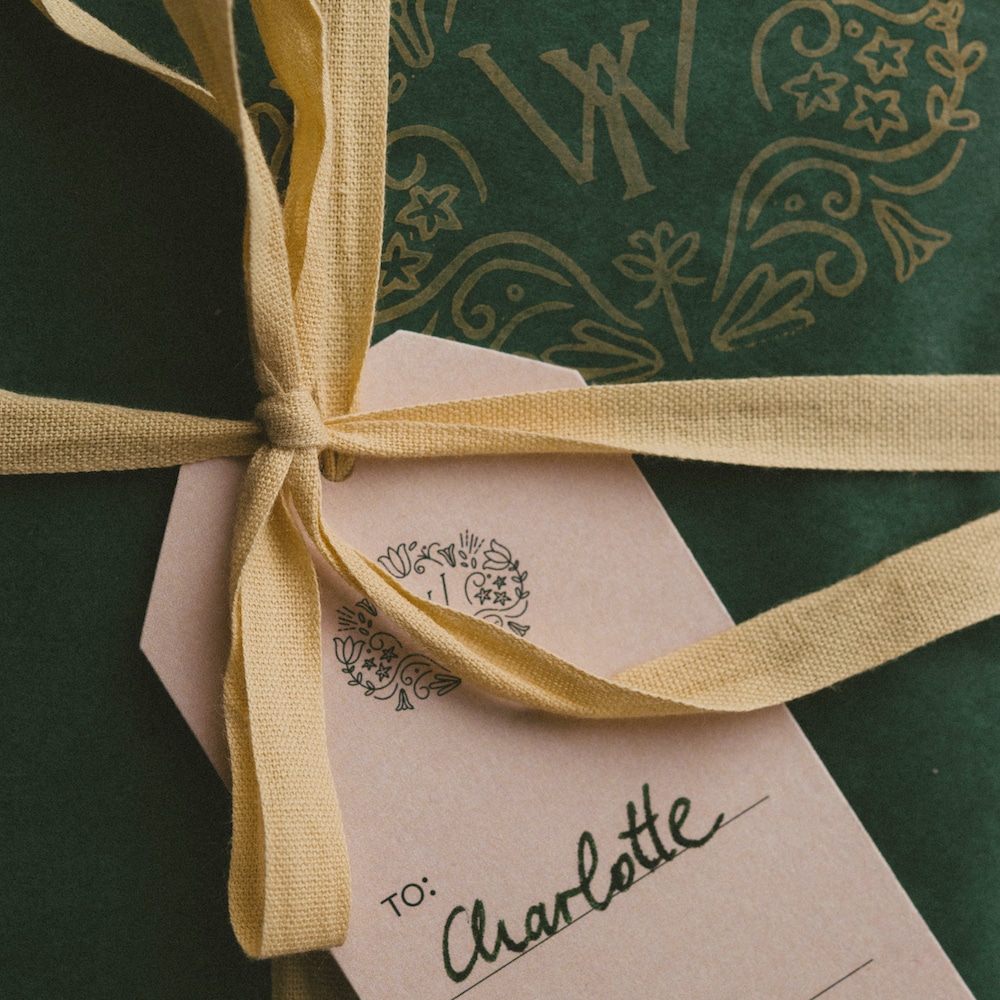 A gift wrapped in green tissue paper stamped with yellow Young W monograms. The gift is packaged with a yellow ribbon and gift tag. The gift tag reads: "To: Charlotte. From: Louise".