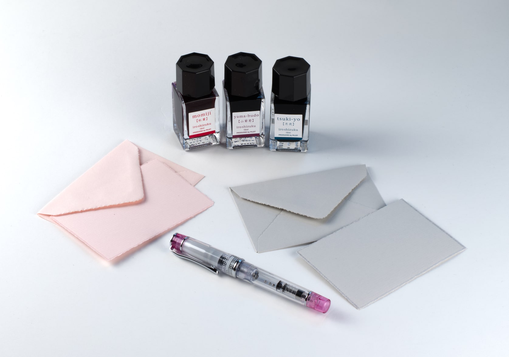 Two mini note card sets with deckled edges in grey and pink, a transparent tinted pink fountain pen and a three-bottle ink set.