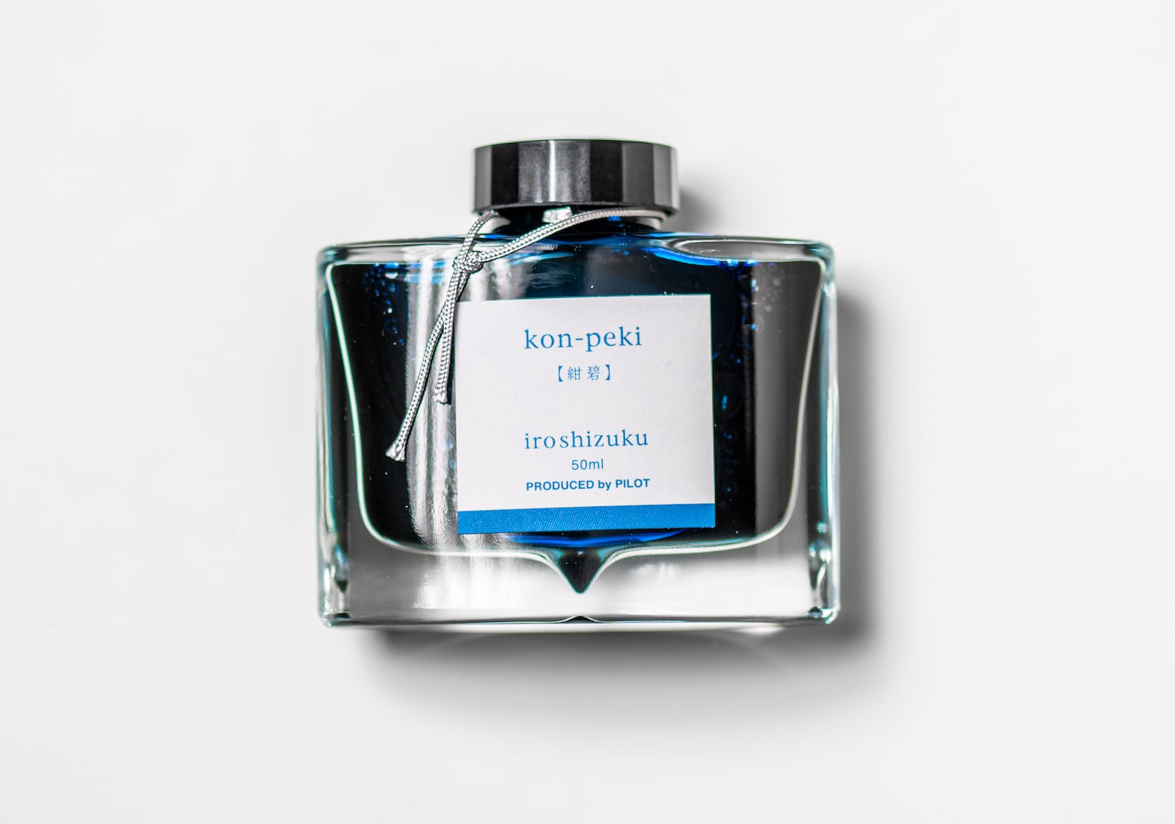 A rectangular glass ink bottle with a silver string tied around the black cap. Square white sticker with blue text that reads: kon-peki. iroshizuku. 50 ml. Produced by Pilot.