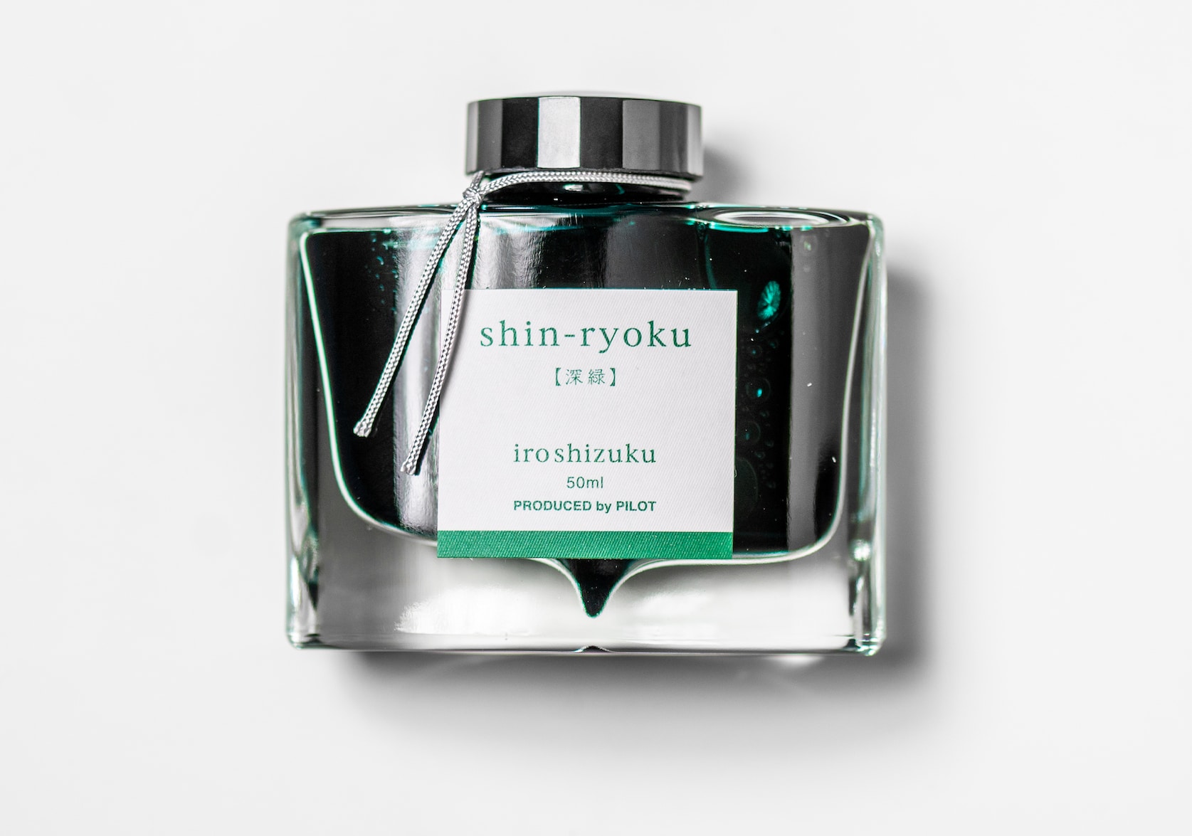 A rectangular glass ink bottle with a silver string tied around the black cap. Square white sticker with green text that reads: shin-ryoku. iroshizuku. 50 ml. Produced by Pilot.