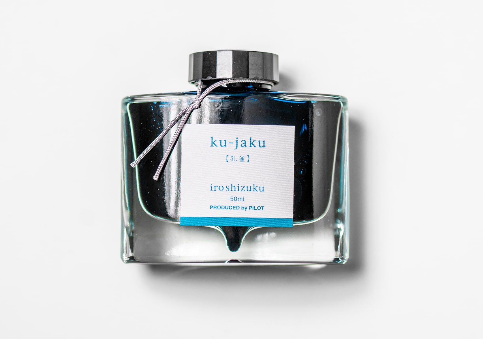 A rectangular glass ink bottle with a silver string tied around the black cap. Square white sticker with blue text that reads: ku-jaku. iroshizuku. 50 ml. Produced by Pilot.