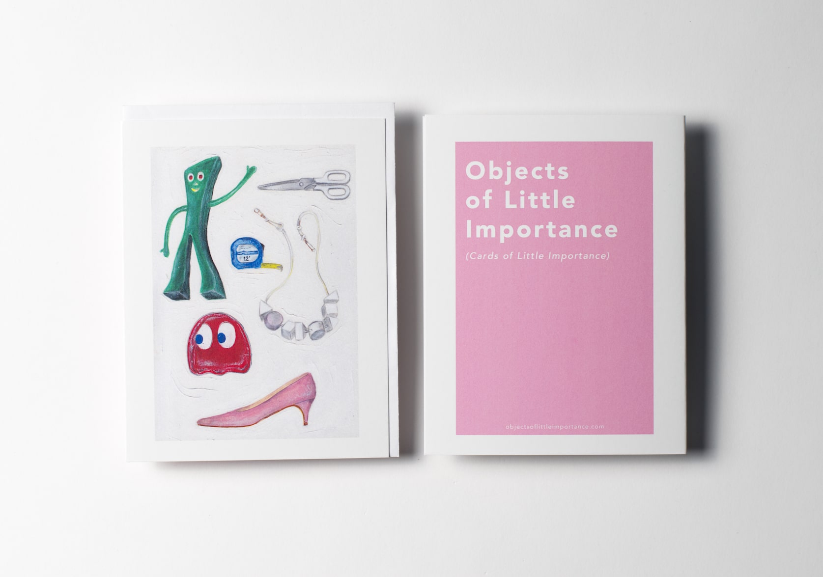 White greeting card featuring a green bendable Gumby figurine, white plastic-handled scissors, a tape measure, a necklace with grey geometric shapes, a red Pac-Man ghost, a pink suede pump. Back of the card is a pink rectangle with white text that reads: Objects of Little Importance (Cards of Little Importance).