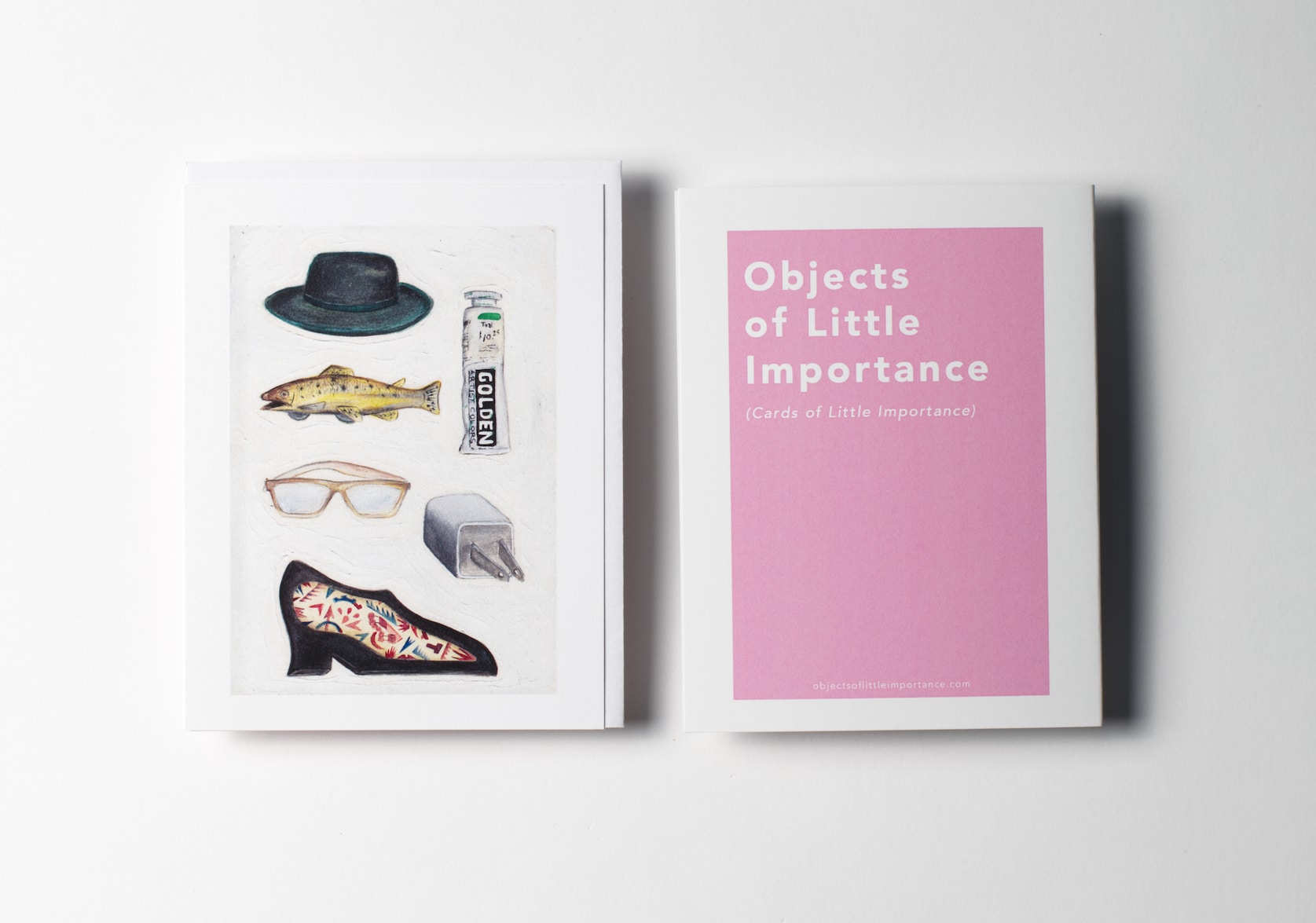 White greeting card featuring paintings of a Loden green hat, a clockwork lithographed tin fish, a tube of acrylic paint, a busted reading glasses, an iPhone cube with a bent prong, a pincushion in the form of a shoe. Back of the card is a pink rectangle with white text that reads: Objects of Little Importance (Cards of Little Importance).