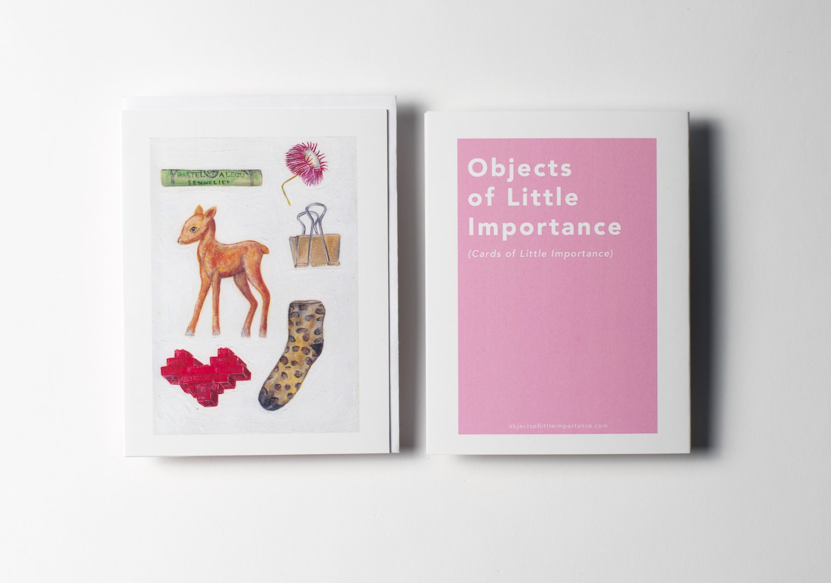White greeting card featuring paintings of a green pastel stick, a dried pink straw flower, a fawn Christmas figurine, a gold paper clip, a red lego heart, a leopard print slipper sock. Back of the card is a pink rectangle with white text that reads: Objects of Little Importance (Cards of Little Importance).