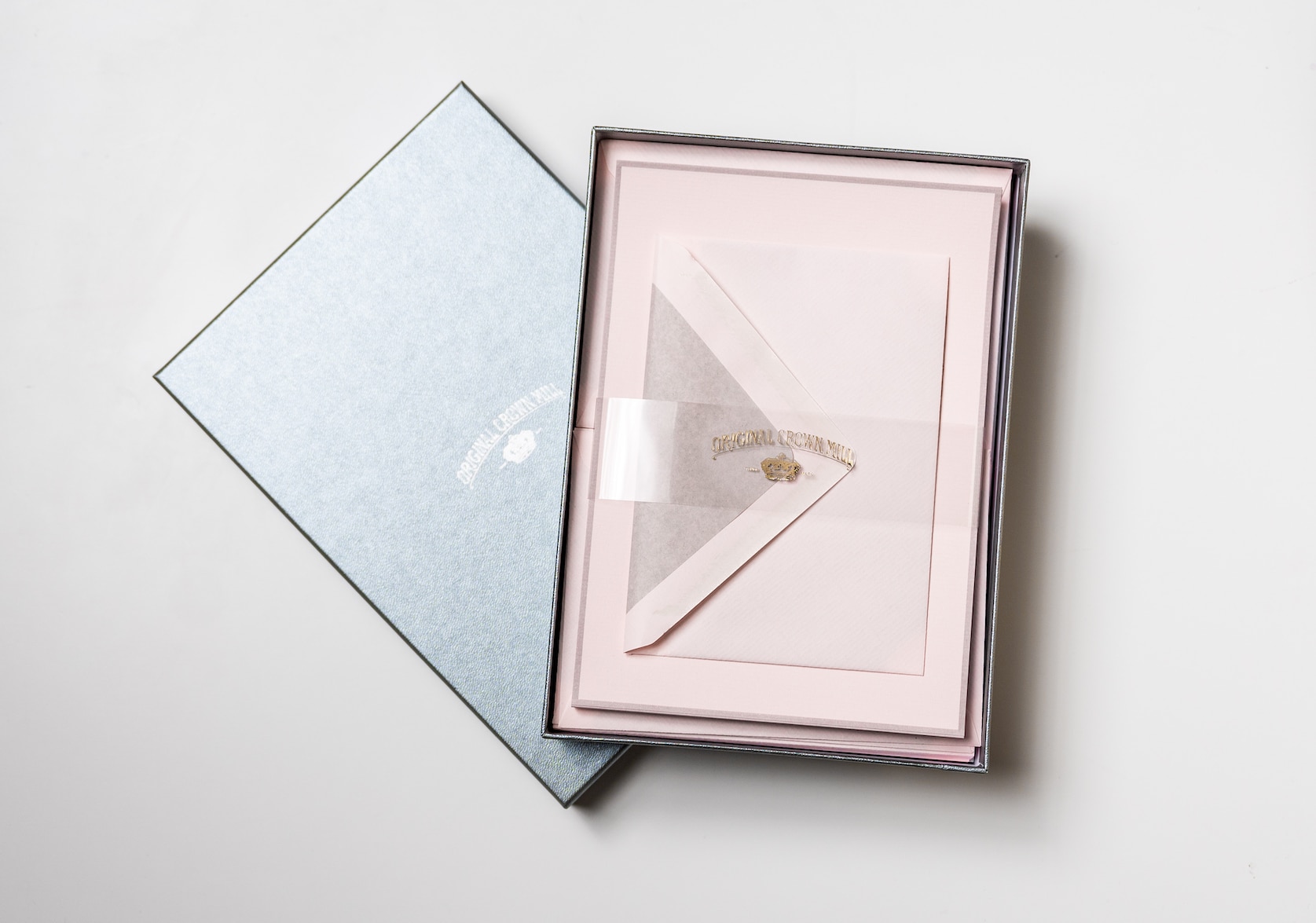Open silver box set of pink stationary with a grey edge. Clear packaging across the box with gold text that reads: Original Crown Mill. Below there is a small gold illustration of a crown.