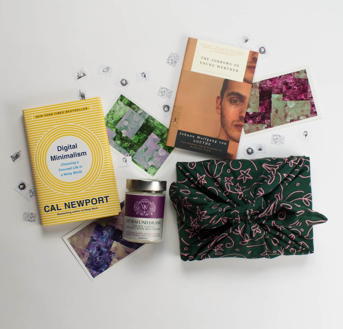 An assortment of products assembled on a white background: the book 'Digital Minimalism', the book 'The Sorrows of Young Werther', a candle with a label that reads 'Sturm und Drang', a gift wrapped in green fabric gift wrap with a floral pattern and a few multi-coloured postcards