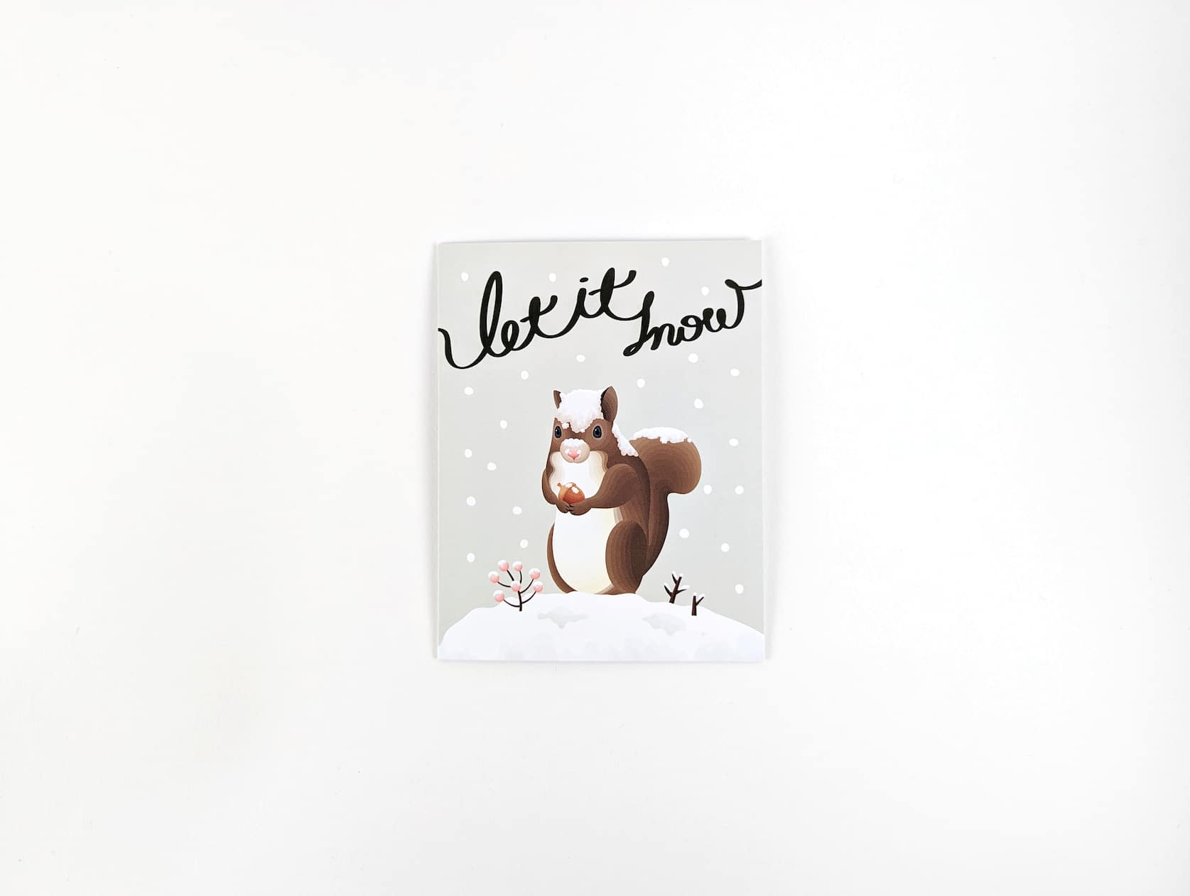 Black cursive text reads: Let it snow. Below a brown squirrel stands in the snow holding an acorn. Beside him a twig of pink berries and twigs stick out of the snow. The background is light grey with snow falling around.