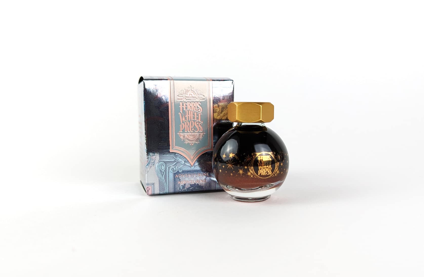 An ornate rose gold, lilac and grey cardboard box with pink text that reads Ferris Wheel Press: Adventurine. Aventurine. Beside the box there is a round glass bottle of ink with a gold lid. On the bottle there is a small gold illustration featuring stars and sparkles.