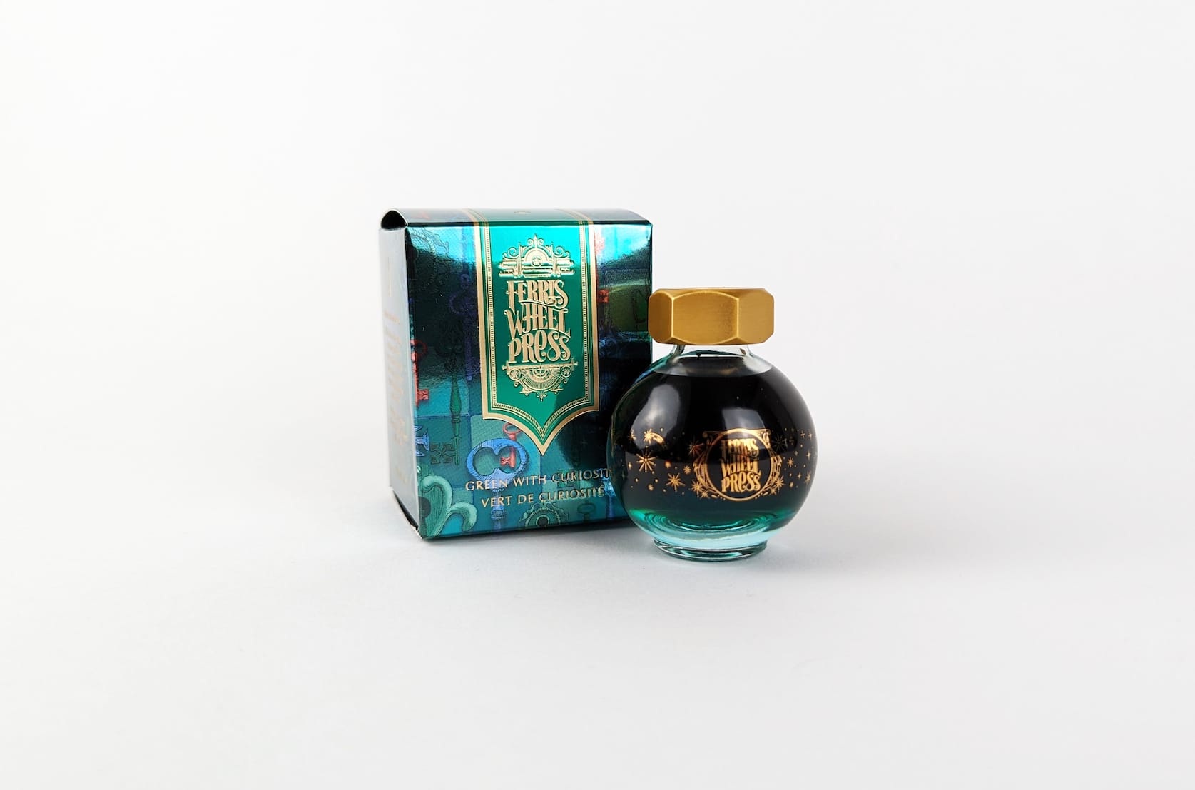 An ornate green, blue, and red cardboard box with gold text that reads Ferris Wheel Press: Green with Curiosity. Vert de Curiosité. Beside the box there is a round glass bottle of ink with a gold lid. On the bottle there is a small gold illustration featuring stars and sparkles.
