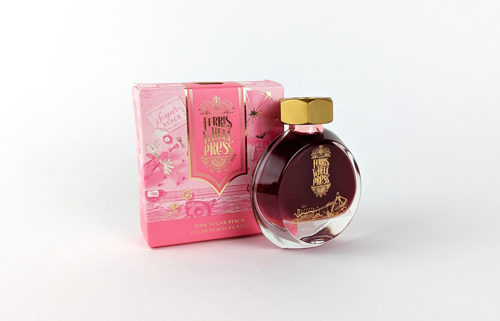 An ornate pink & gold cardboard box with gold text that reads Ferris Wheel Press: Pink Sugar Beach. Sugar Beach en Rose. Beside the box there is a round glass bottle of ink with a gold lid. On the bottle there is a small gold illustration featuring a merry-go-round and circus tent.
