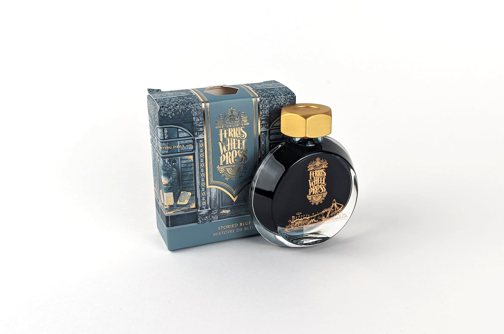 An ornate blue & gold cardboard box with gold text that reads Ferris Wheel Press: Storied Blue. Histoire de Bleu. Beside the box there is a round glass bottle of ink with a gold lid. On the bottle there is a small gold illustration featuring a merry-go-round and circus tent.