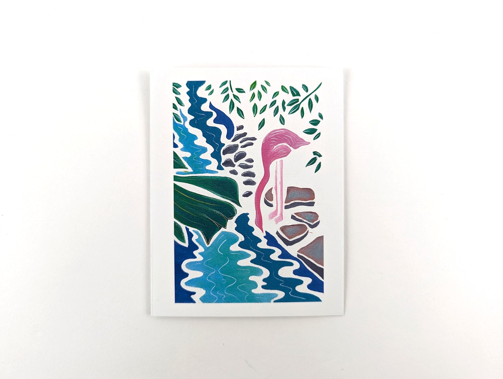 White card with a painted pink flamingo drinking at a blue stream. Grey rocks line the water's edge and there is green leaves hanging above