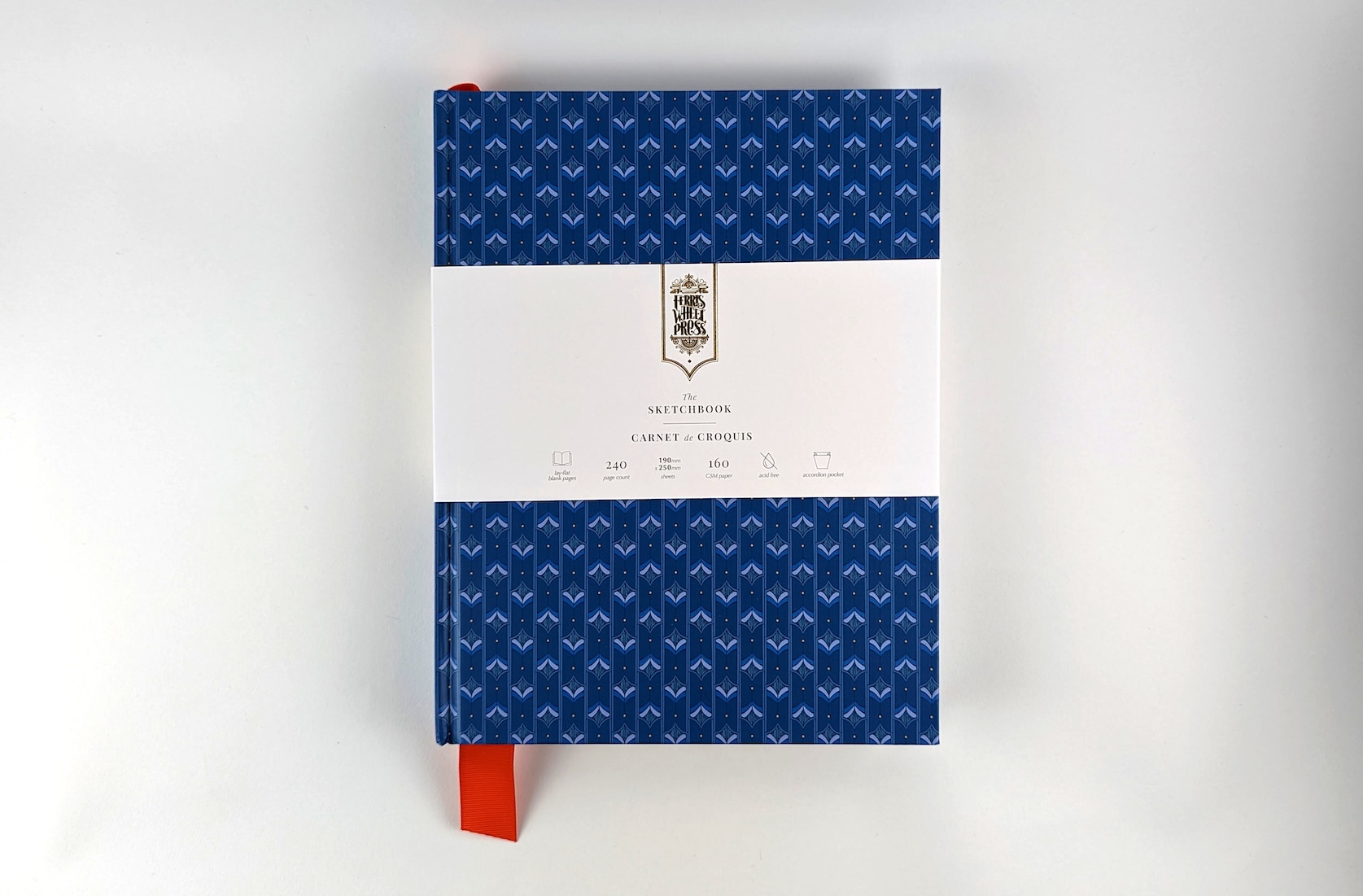 A blue sketchbook covered in paper with a repeating damask pattern featuring a red fabric ribbon sticking out of the bottom. Gold text on the notebook's packaging reads: Ferris Wheel Press logo. The Sketchbook. Carnet de Croquis.