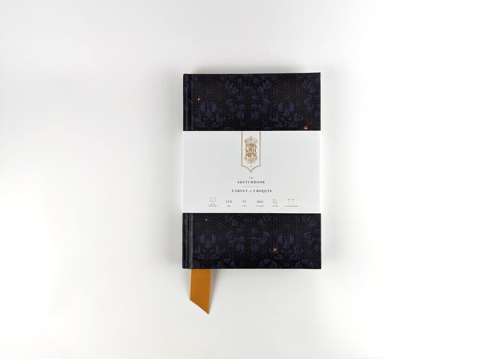A dark blue sketchbook covered in paper with a repeating damask pattern featuring a gold fabric ribbon sticking out of the bottom. Gold text on the notebook's packaging reads: Ferris Wheel Press logo. The Sketchbook. Carnet de Croquis.