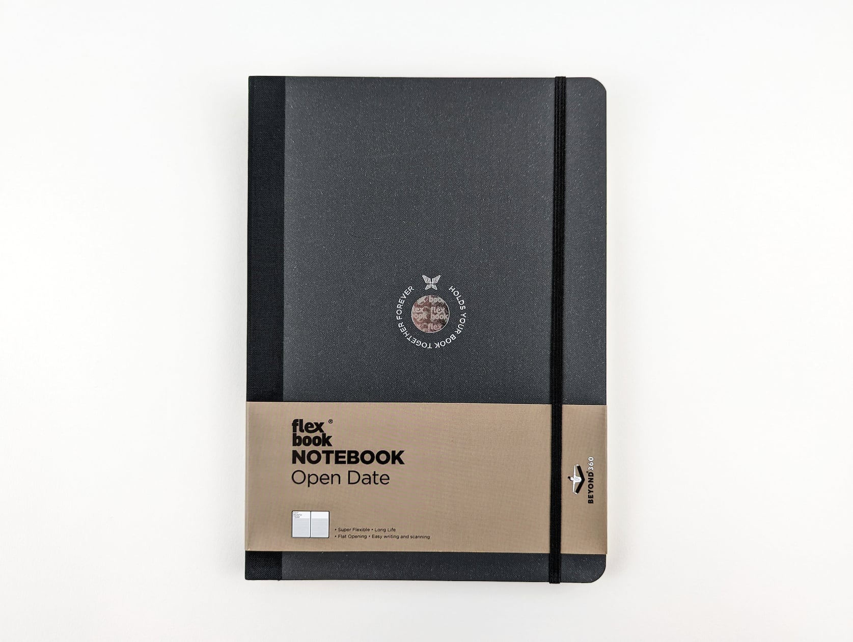 A black notebook with a black fabric spine and elastic band closure. A circular holographic logo is featured in the center with a butterfly and silver text wrapping around that reads: Holds your book together forever. Light brown paper packaging is located across the bottom half of the notebook with black text that reads: flexbook Notebook Open Date. Super Flexible. Long Life. Flat Opening. Easy Writing and Scanning. Beyond 360.