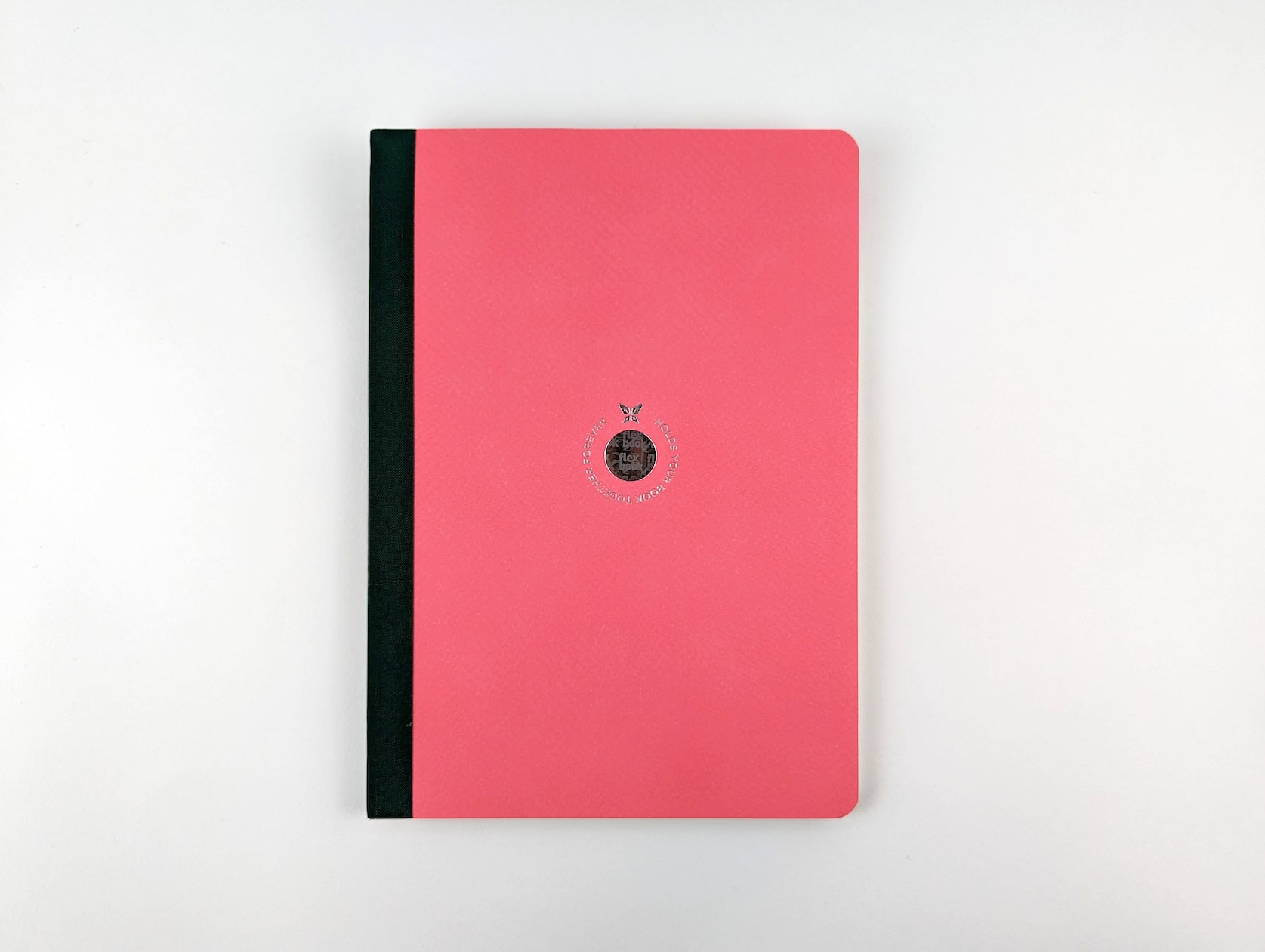 A notebook with pink eco paper and a black linen spine. A circular holographic logo is featured in the center with a butterfly and silver text wrapping around that reads: Holds your book together forever.