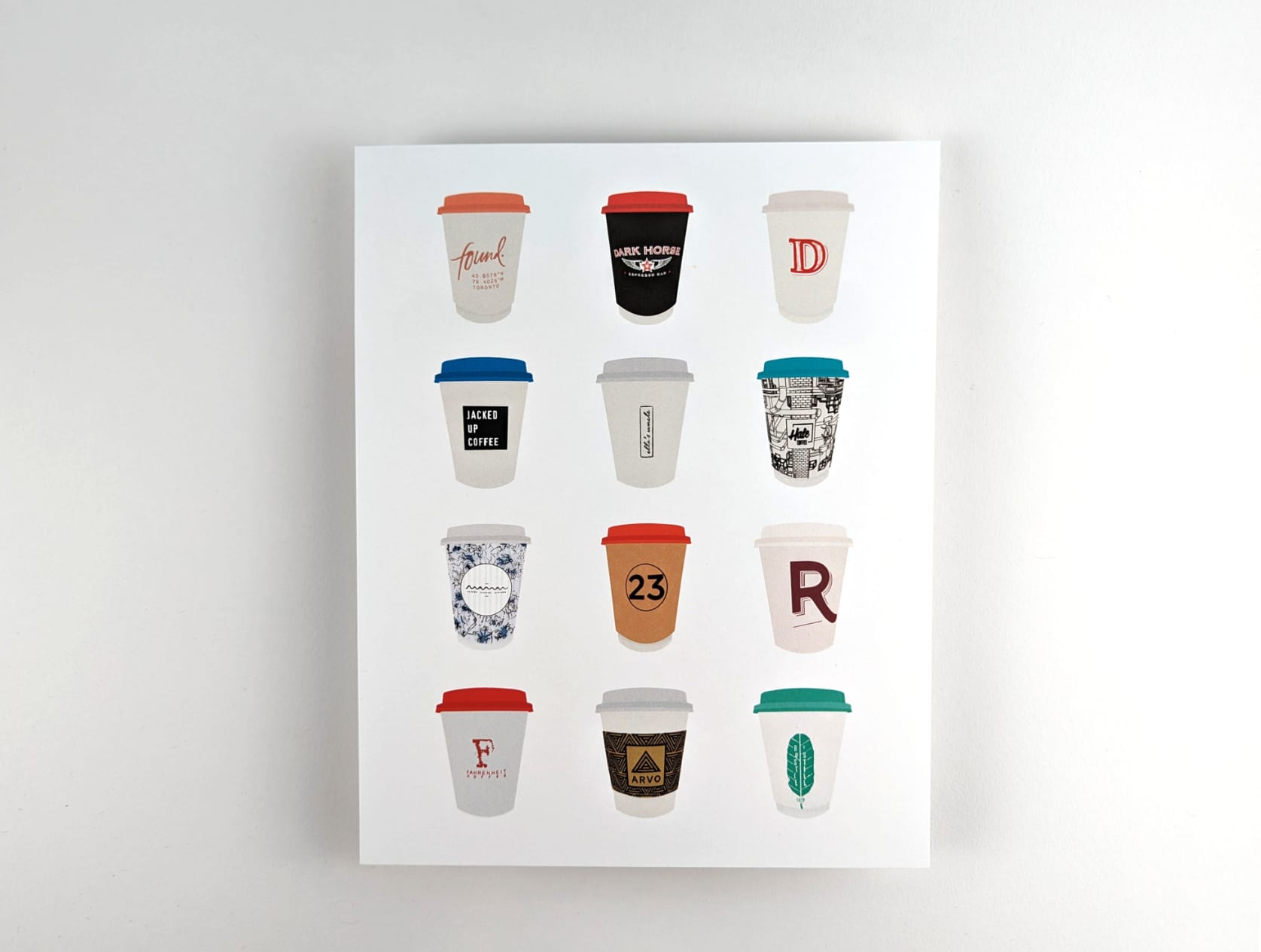 Print featuring illustrations of twelve coffee to-go cups from Toronto cafés against a white background. The twelve coffee cups are arranged in 4 rows with 3 cups in each row.