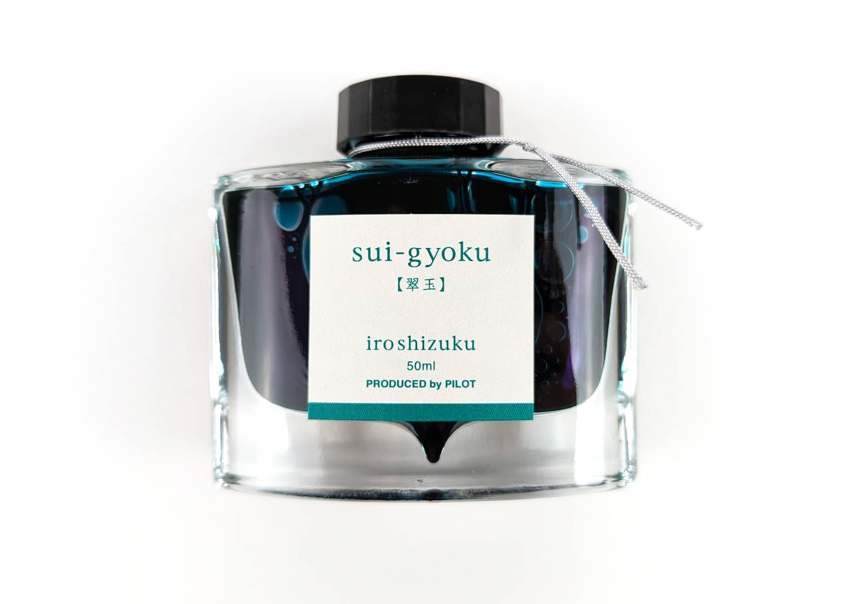 A rectangular glass ink bottle with a silver string tied around its neck. Square sticker with green text that reads: sui-gyoku. iroshizuku. 50 ml. Produced by Pilot.
