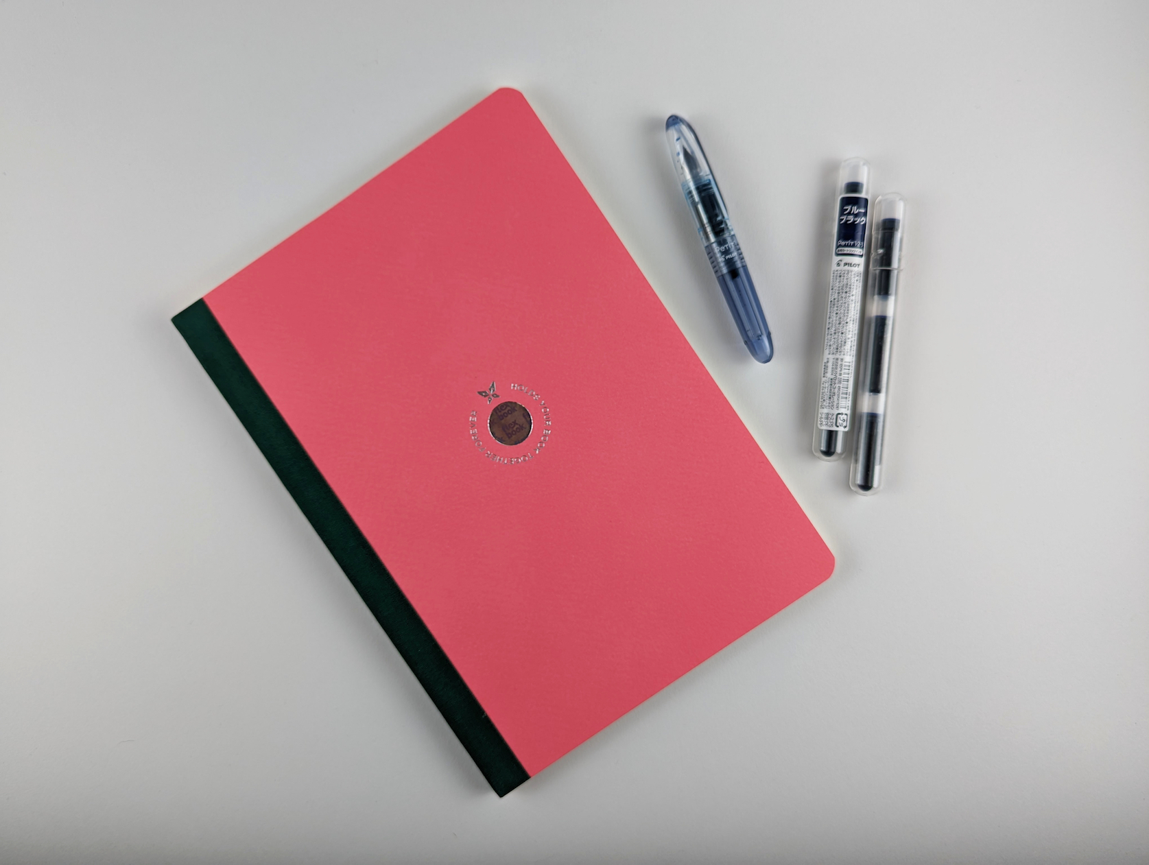 Bundle of items on a white background. Items include: pink notebook, blue-black fountain pen and two packs of blue black fountain pen ink cartridges.