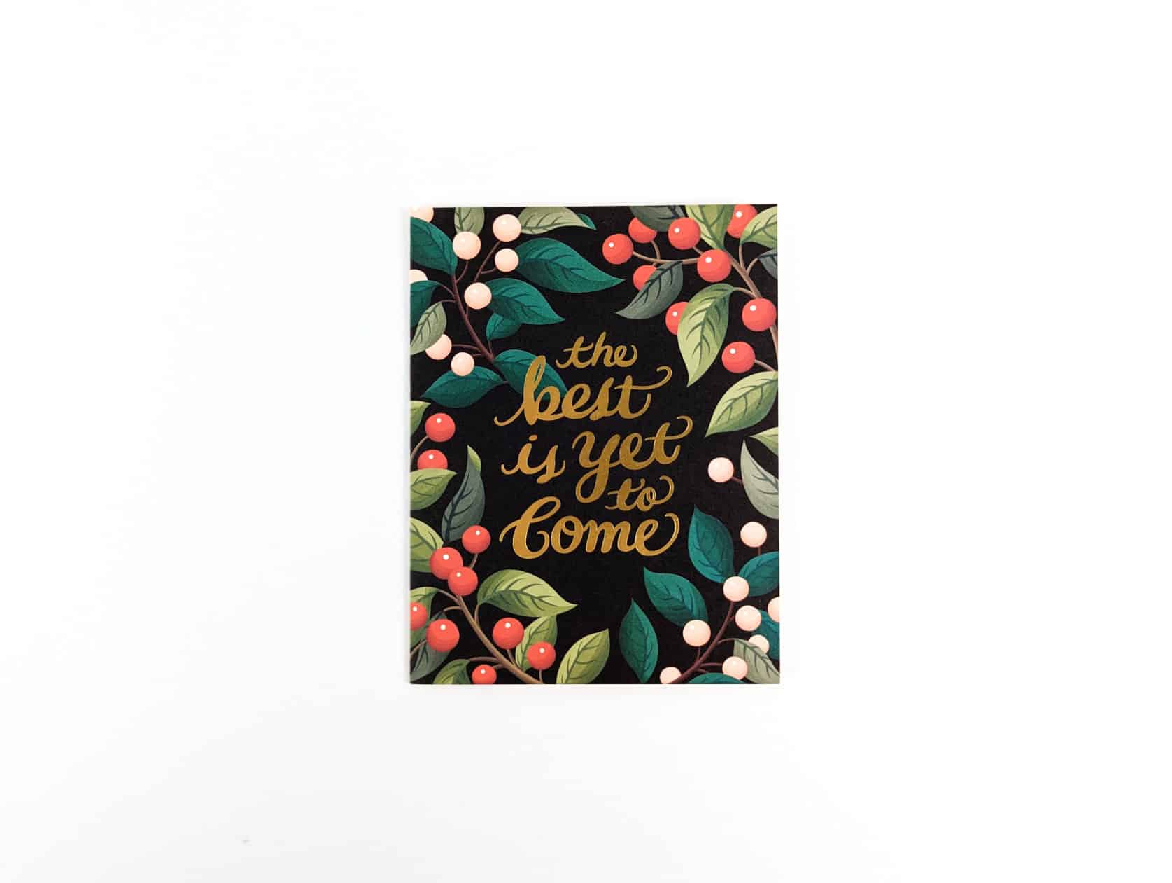 Black card with gold cursive text in the centre that reads: the best is yet to come. Red & pink wild berries and greenery surround the text.