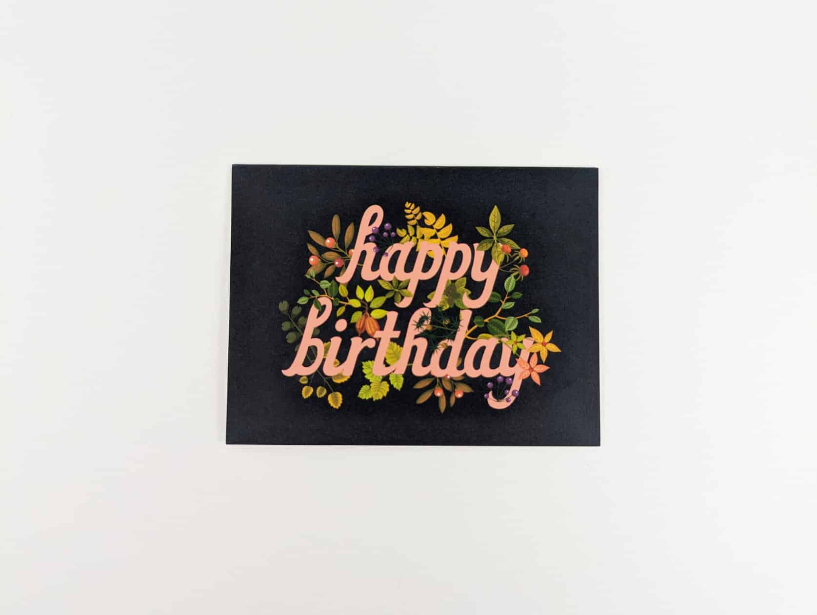 Navy horizontal card with pink cursive writing that reads: happy birthday. Sticking out from behind the text are yellow & green leaves, pink & yellow flowers, red & purple berries.