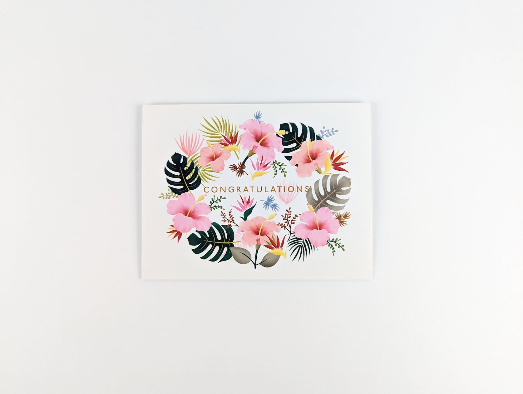 Cream horizontal card with gold text in the centre that reads: CONGRATULATIONS. Hibiscus flowers, green & grey monstera leaves and blue, green and brown leaves surround the text in a ring.