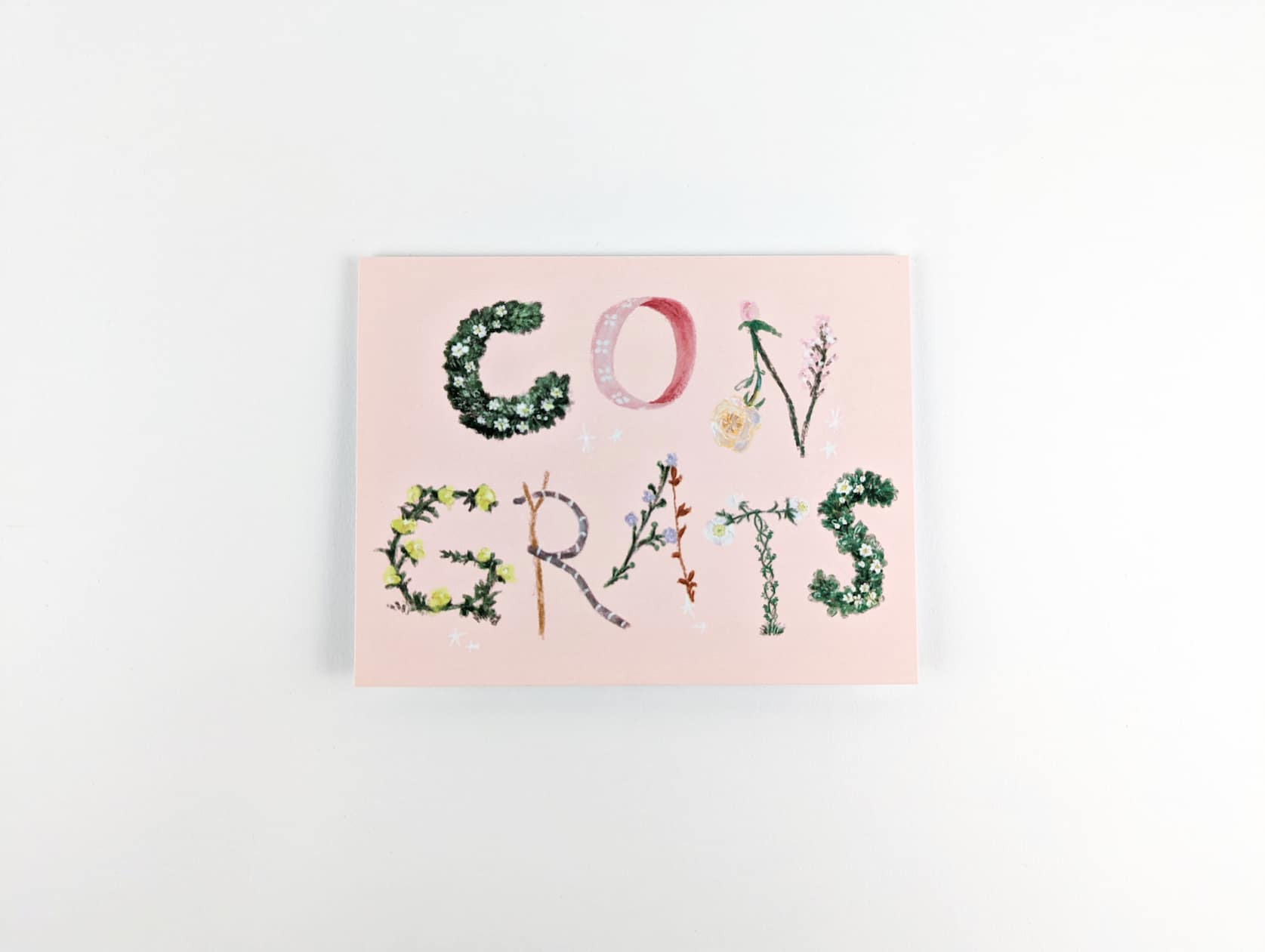 Pink horizontal card with the text: CONGRATS. Congrats is spelling out in floral lettering with shrubbery, ribbon, flowers, leaves & twigs.