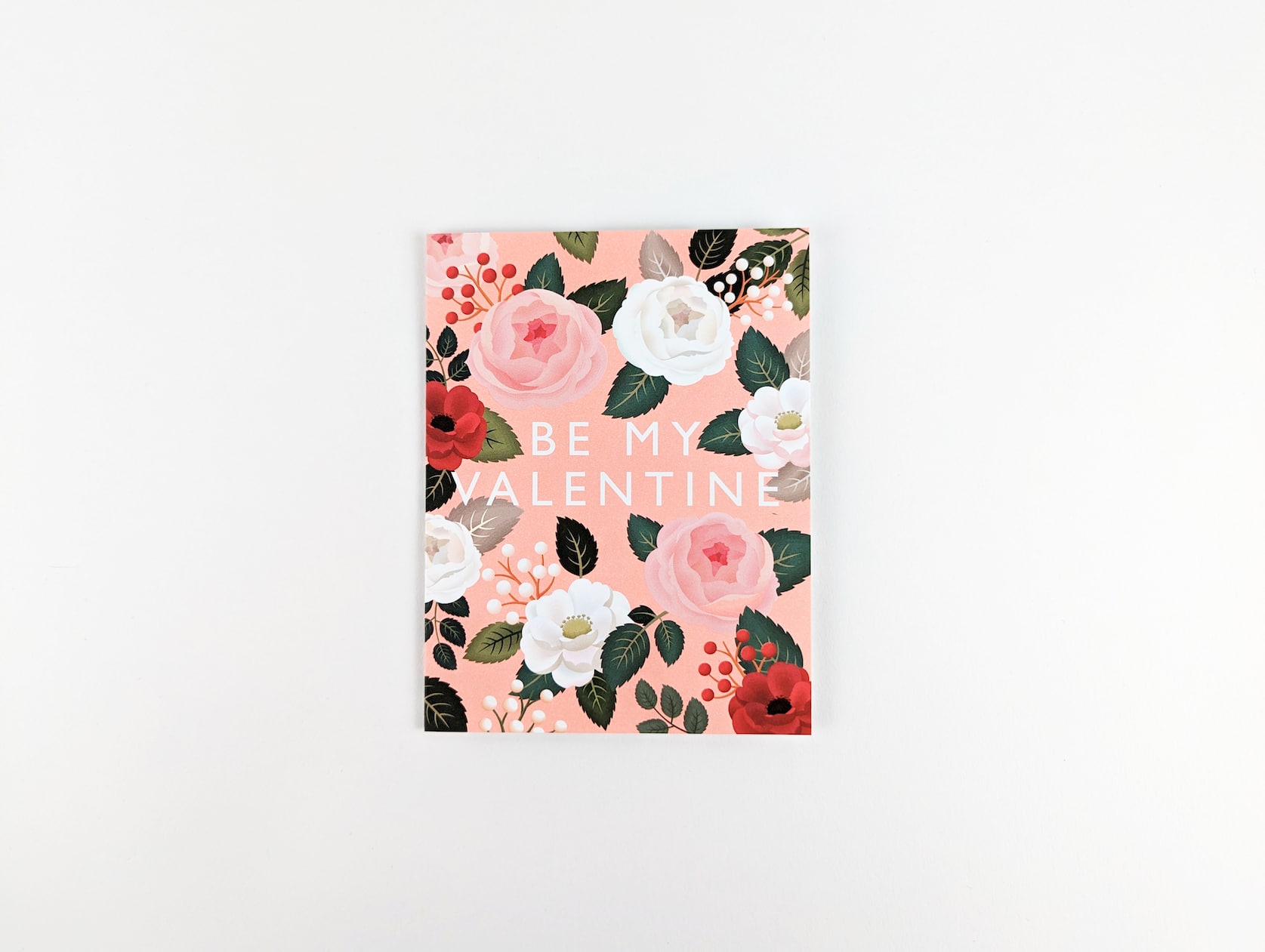 Pink card with white text in the centre that reads: BE MY VALENTINE. White, pink and red roses with green leaves and matching berries appear behind the text.