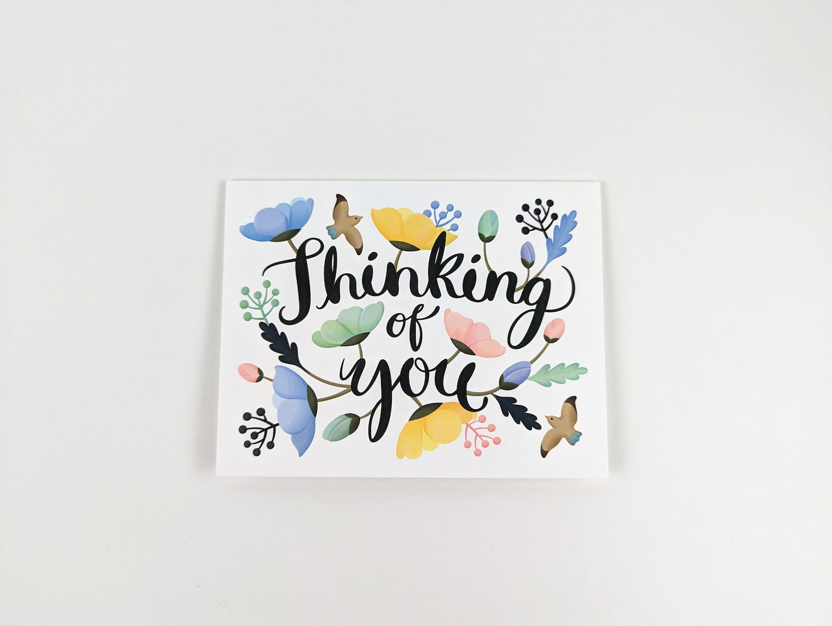 White horizontal card with black cursive text in the centre that reads: Thinking of you. A variety of pastel blue, yellow, green and pink poppies, sprigs and two brown songbirds are featured around the text.