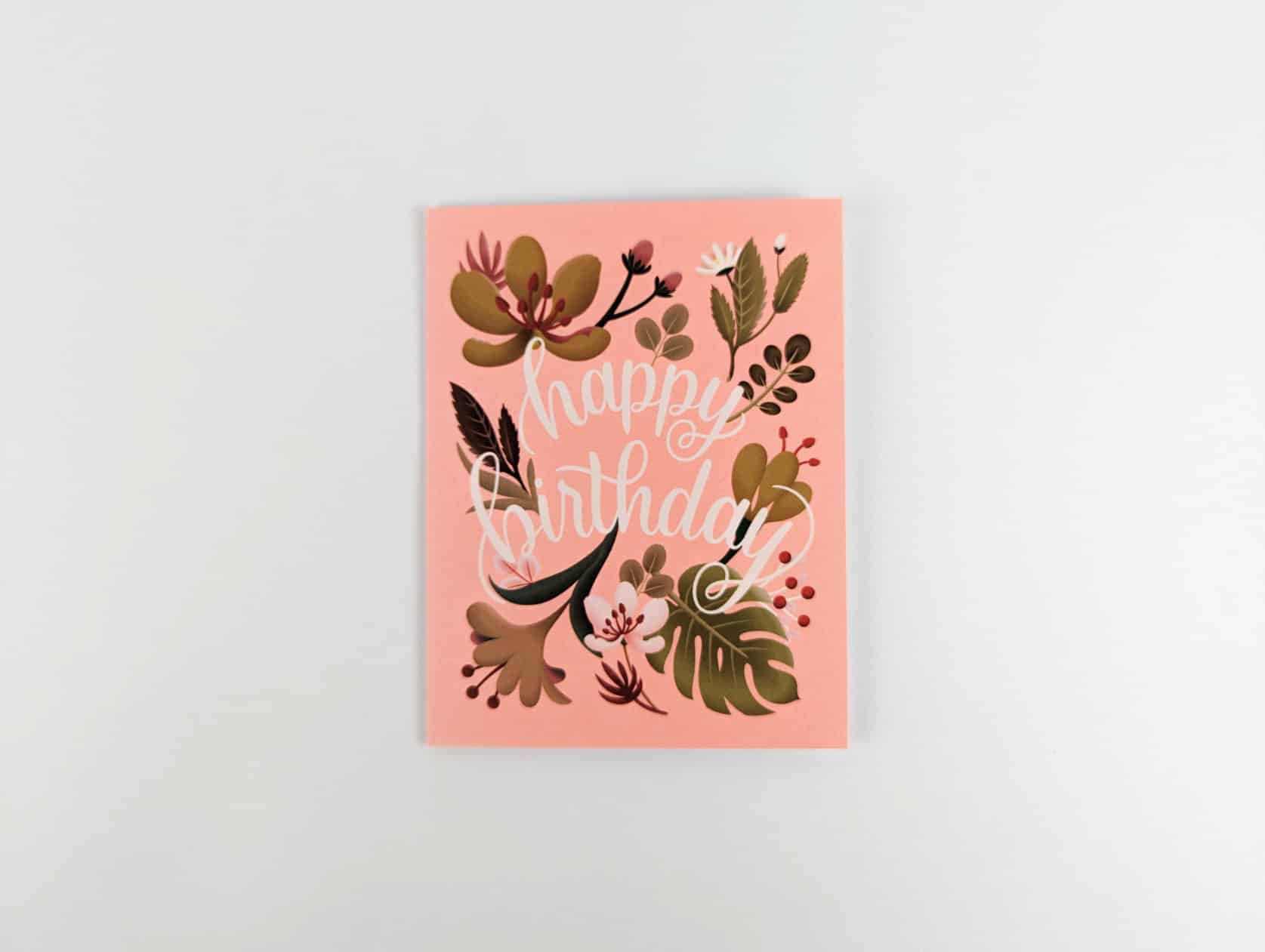 Coral card with white cursive text in the centre that reads: happy birthday. A variety of pink, white and green tropical flowers and foliage are featured around the text.