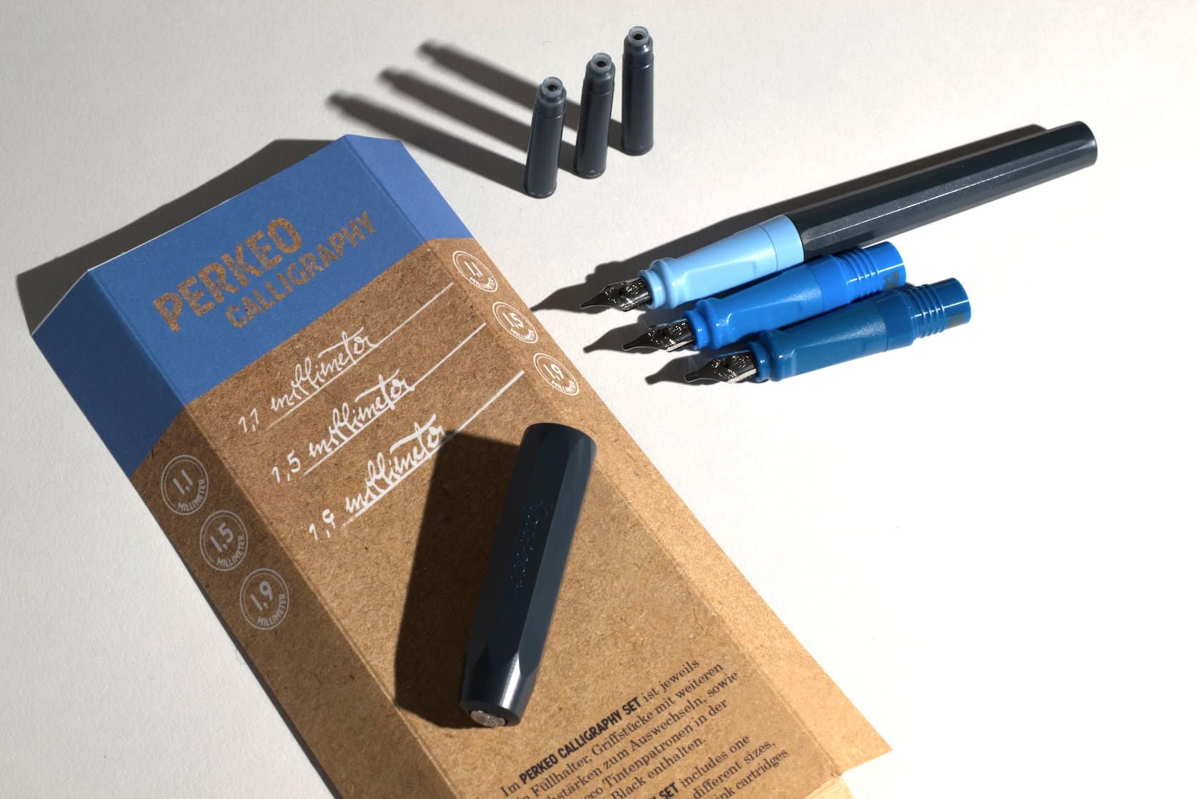 A biue/dark blue fountain pen lies on a white surface. Next to it are two fountain pen front parts with slightly broader nibs and three ink cartridges. A portion of the product packaging is visible. Text reads: Perkeo Calligraphy. It shows writing samples of the three included nib sizes. The nib sizes are 1.1 mm, 1.5 mm and 1.9 mm.