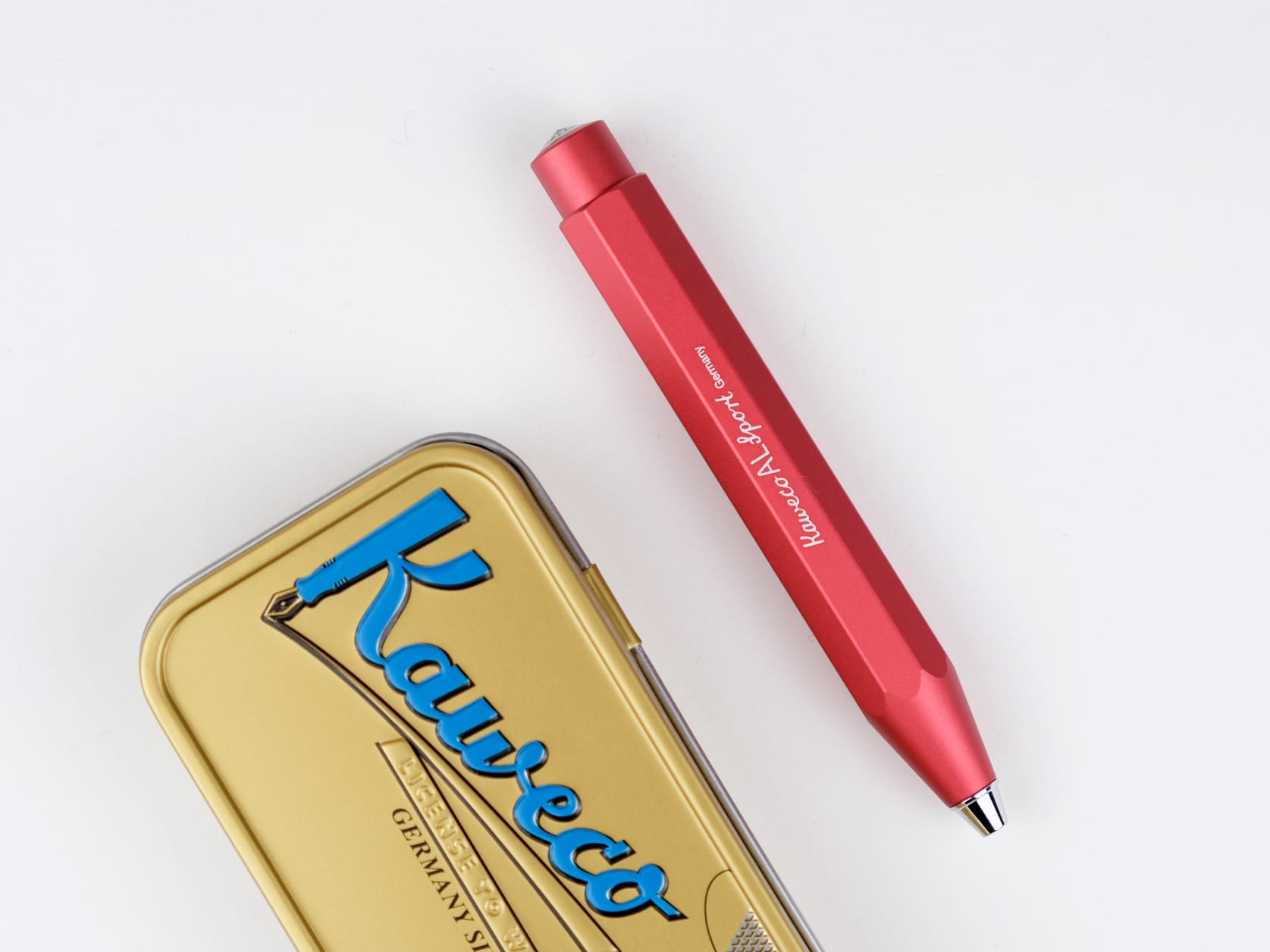 A red aluminum ballpoint pen lying beside its golden tin which has Kaweco written on it in big blue letters