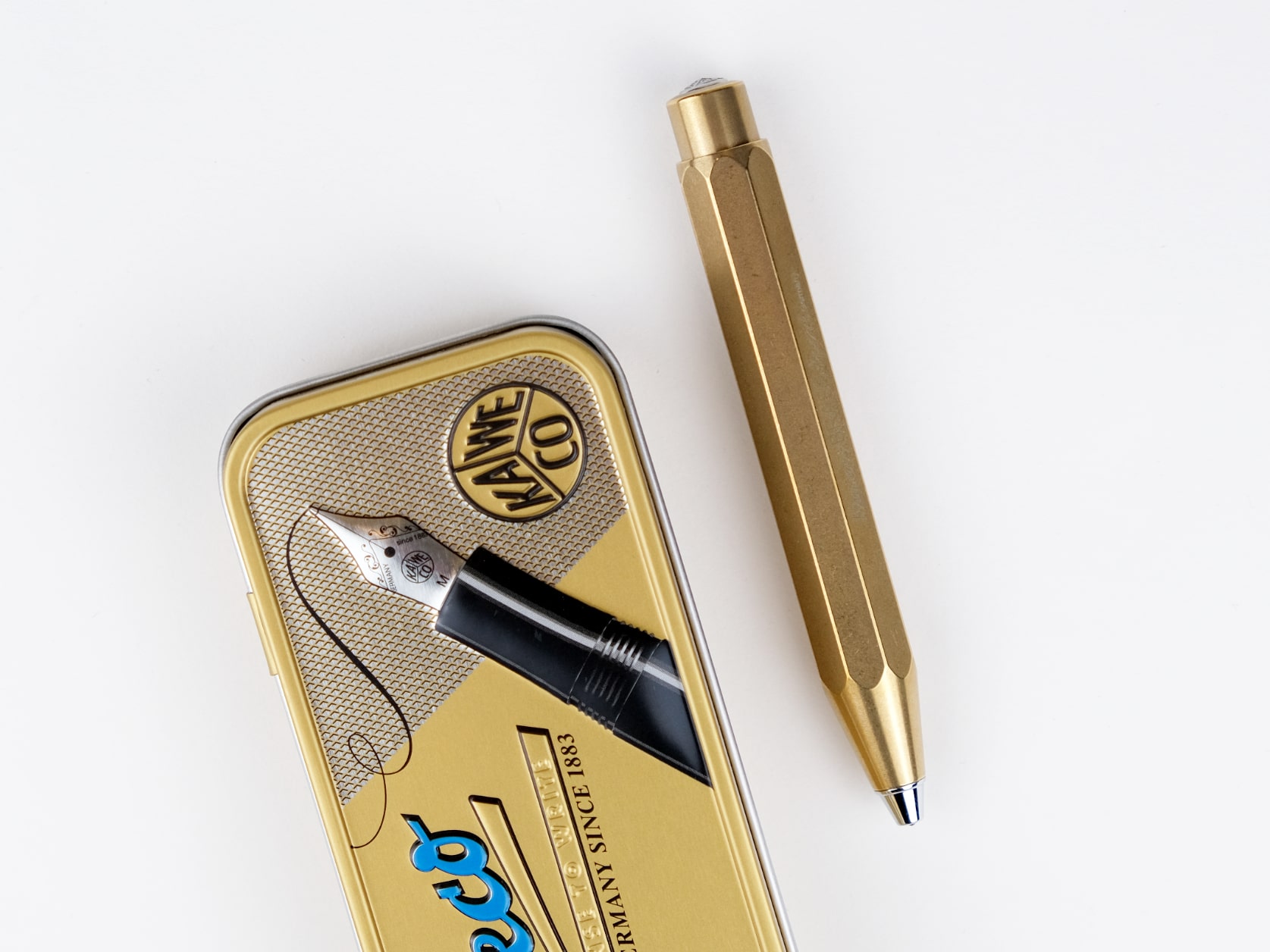 A brass ballpoint pen lying beside its golden tin